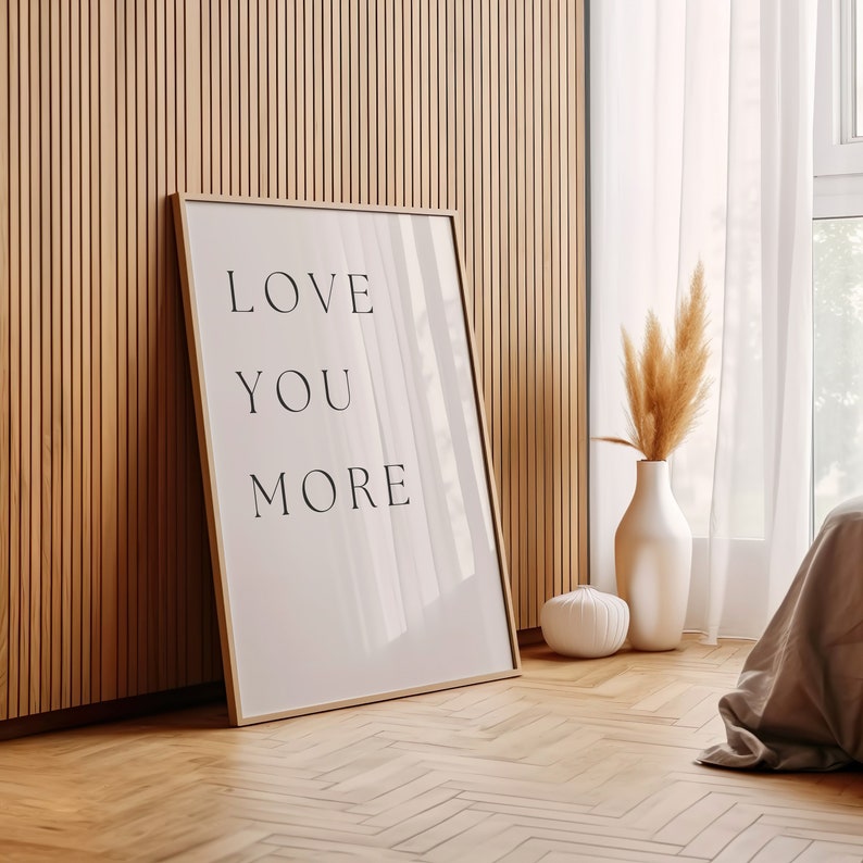 K&ouml;nnte beinhalten: Ein gerahmter Druck mit den Worten "LOVE YOU MORE" in schwarzer Schrift auf wei&szlig;em Hintergrund. Der Druck lehnt an einer Holzpaneelwand mit einer Vase mit getrocknetem Pampasgras und einem wei&szlig;en Dekorationsobjekt im Vordergrund.
