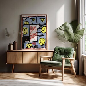 May include: A vintage poster with a colorful abstract design featuring tennis balls and the text "Roland Garros 1988". The poster is framed and hanging on a wall above a wooden cabinet and a green armchair.