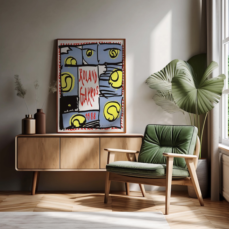 May include: A vintage poster with a colorful abstract design featuring tennis balls and the text "Roland Garros 1988". The poster is framed and hanging on a wall above a wooden cabinet and a green armchair.