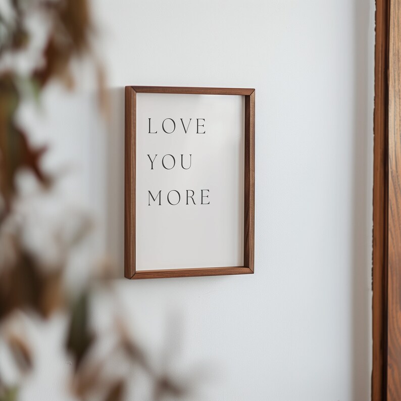 K&ouml;nnte beinhalten: Ein gerahmter Druck mit dem Text "LOVE YOU MORE" in einer einfachen, modernen Schriftart. Der Rahmen ist aus hellem braunem Holz.
