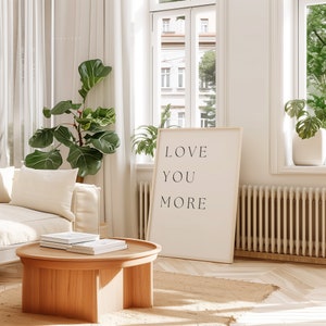 K&ouml;nnte beinhalten: Ein wei&szlig;er gerahmter Poster mit dem Text "LOVE YOU MORE" h&auml;ngt an einer wei&szlig;en Wand in einem Wohnzimmer. Der Poster lehnt an einem wei&szlig;en Heizk&ouml;rper. Eine gr&uuml;ne Pflanze steht in der Ecke des Raumes.