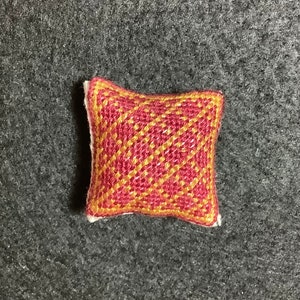 Puede incluir: Una pequeña almohada decorativa cuadrada con un patrón tejido rojo y dorado. La almohada tiene un borde blanco y está sobre un fondo gris. El diseño tejido crea un patrón de diamantes.