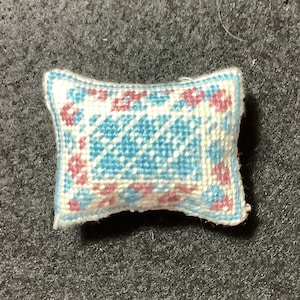 Puede incluir: Una pequeña almohada blanca con un diseño bordado azul y rosa. La almohada es rectangular y tiene una forma ligeramente redondeada.