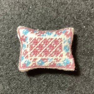 Puede incluir: Una pequeña almohada blanca con un diseño bordado en rosa y azul. La almohada tiene una forma cuadrada con esquinas redondeadas.