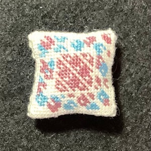 Puede incluir: Una pequeña almohada blanca con un diseño bordado en rosa y azul. La almohada es de forma cuadrada y tiene una tela suave y texturizada.