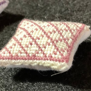 Puede incluir: Pequeño cojín decorativo con base blanca y diseño geométrico rosa y blanco. El cojín tiene un aspecto suave y texturizado y un borde festoneado. El diseño es un patrón de diamantes repetido.