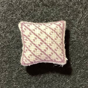 Puede incluir: Una pequeña almohada cuadrada con fondo blanco y un patrón diagonal de rayas moradas.