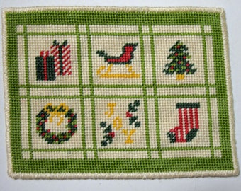 May include: A green and white cross-stitch sampler with a Christmas theme. The sampler features six squares with different Christmas motifs, including presents, a sleigh, a Christmas tree, a wreath, holly, and a stocking.