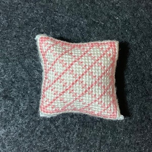 Puede incluir: Una pequeña almohada blanca con un patrón de rombos rosa y blanco. La almohada está hecha de una tela suave y tejida.