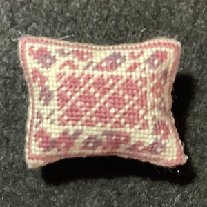 Puede incluir: Una pequeña almohada rosa y blanca con un patrón floral. La almohada está hecha de una tela suave y texturizada y tiene una forma cuadrada.