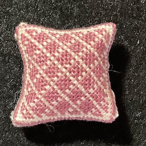 Puede incluir: Una pequeña almohada decorativa cuadrada con un patrón de pata de gallo rosa y blanco. La almohada en miniatura está hecha de tela tejida y tiene una apariencia suave y texturizada. La almohada es de color rosa claro con detalles blancos.