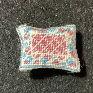 Puede incluir: Una pequeña almohada blanca con un diseño bordado en rosa y azul. La almohada es rectangular y tiene una forma ligeramente redondeada.