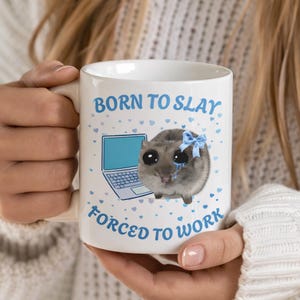 Peut inclure: Mug en céramique blanche avec l'inscription bleue « BORN TO SLAY FORCED TO WORK ». La tasse présente une illustration de hamster qui pleure avec un nœud bleu et un ordinateur portable. Petits cœurs bleus éparpillés.