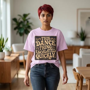 Peut inclure: Un t-shirt lilas avec le texte "THIS DANCE THING ESCALATED QUICKLY" dans un design vintage. Le texte est marron foncé avec des accents dorés. La personne porte un jean bleu foncé.