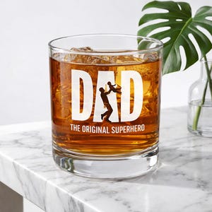 Peut inclure: Un verre à whisky transparent rempli de liquide ambré et de glaçons. Le verre porte l'inscription "DAD" en grandes lettres blanches, avec une silhouette d'un père et d'un enfant. En dessous, le texte dit "THE ORIGINAL SUPERHERO."