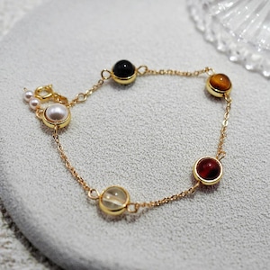 Może przedstawiać: Złota bransoletka z łańcuszkiem i pięcioma okrągłymi koralikami z kamieni szlachetnych. Koraliki są czarne, białe, brązowe, przezroczyste i czerwone.