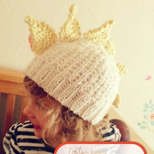 May include: A child wearing a white knitted hat with a yellow sun design. The hat has a pointed top with five points. The hat is a PDF pattern for sale.