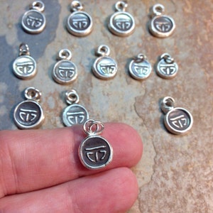 May include: A collection of silver charms with a stylized symbol, some with a pearlescent finish. The charms are round and have a loop at the top for attaching to a necklace or bracelet.