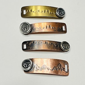 May include: Four metal tags with a mountain range design and a circular logo. The tags are in different shades of copper and brass.