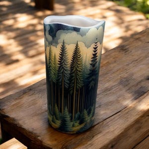 May include: A tall, cylindrical candle with a forest scene printed on it. The candle features a blue and green color scheme with a depiction of a forest with tall trees and a cloudy sky.