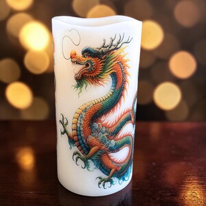 May include: A cylindrical white pillar candle featuring a detailed, colorful illustration of a dragon with orange, green, and blue scales. The dragon is depicted in a winding pose with visible claws, whiskers, and horns against the smooth surface.