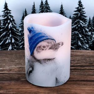 May include: A white candle with a watercolor image of a penguin wearing a blue knit cap. The penguin is set against a snowy forest backdrop. The candle is cylindrical and has a slightly curved top.