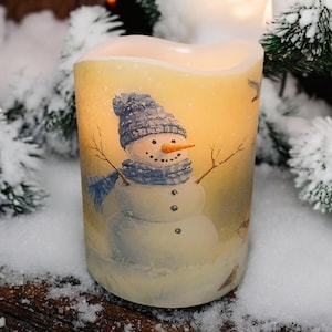 May include: A battery-operated snowman candle with a warm, flickering glow. The candle features a painted snowman wearing a blue hat and scarf, with twig arms and a carrot nose. The candle is surrounded by artificial snow and evergreen branches.