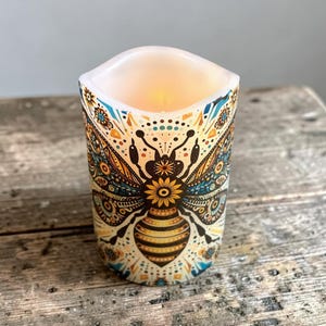 May include: A cylindrical LED candle featuring a detailed graphic of a bee with intricate wings. The design incorporates a warm color palette of yellow, orange, and brown, accented with teal and black patterns. The top edge has a wavy, uneven rim.
