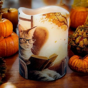 May include: A white, battery-operated candle with a cat wearing glasses reading a book. The candle has a fall theme with pumpkins and a tree with orange leaves. The cat is sitting in a chair.