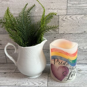 May include: A white ceramic pitcher with a handle holds green fern-like plants. Beside it is a battery-operated candle with a rainbow design and the words "Pride in the world of Love" on the front.