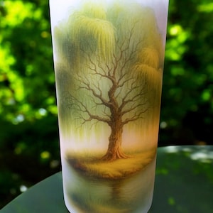 May include: A tall, cylindrical glass with a hand-painted landscape featuring a weeping willow tree. The artwork depicts a tree with a brown trunk and branches, surrounded by green foliage and a reflection in the water. The glass has a frosted appearance.