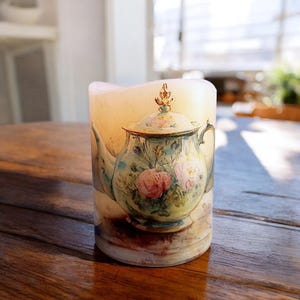 May include: A white pillar candle with a floral design of a teapot with pink and green flowers. The candle is on a wooden surface.