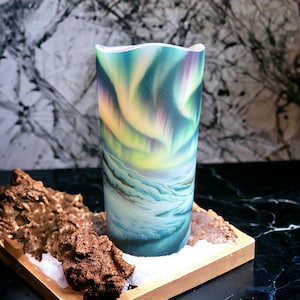May include: A cylindrical glass candle with a colorful aurora borealis design. The candle is sitting on a wooden surface with a piece of driftwood and white snow.
