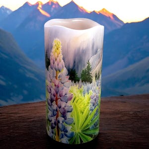 May include: A cylindrical candle with a watercolor painting of a mountain landscape and wildflowers. The candle features a light blue, purple, and green floral design with a mountain range in the background. The candle is approximately 6 inches tall.