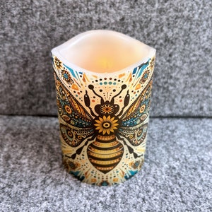 May include: A white pillar candle with a colorful bee design. The bee has a yellow and black striped body and a sunflower for a head. The candle is lit and glowing.