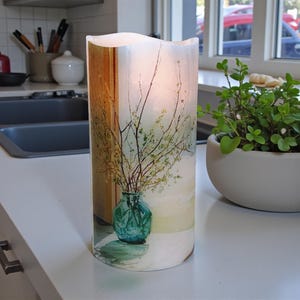 May include: A cylindrical LED candle with a watercolor design. The design features a teal vase with branches and small green leaves. The candle is on a white countertop near a window and a small potted plant.