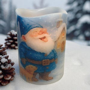May include: A battery-operated candle with a whimsical illustration of a smiling gnome in a blue coat and hat, accompanied by a small dog. The design includes a snowy background and a small house, evoking a winter theme.