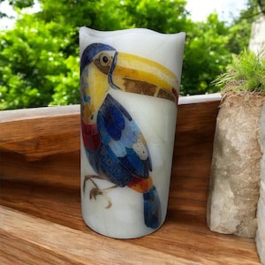 May include: A white candle with a colorful toucan design. The toucan features blue, yellow, red, and brown details. The candle is cylindrical with a slightly wavy top, and is set against a natural wood surface.