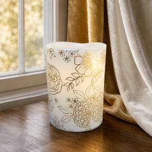 May include: A cylindrical white LED candle featuring a gold-colored floral line art pattern. The design includes various blossoms, leaves, and stems covering the exterior surface. The top edge of the candle has a slightly uneven, wavy rim.