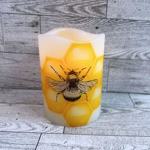 May include: A white pillar candle with a yellow and black bee design on a honeycomb pattern. The candle is lit and glowing.