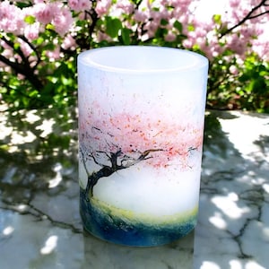 May include: A white pillar candle with a watercolor design of a pink cherry blossom tree. The candle is on a marble surface.