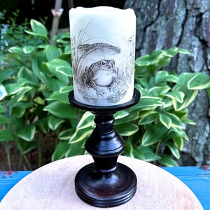 May include: A white pillar candle with a black and white illustration of a frog sitting on a lily pad. The candle is on a black wooden pedestal.