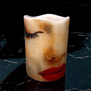 May include: A white pillar candle with a printed image of a woman's face with closed eyes and red lips. The candle has a textured surface.