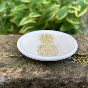 May include: A white ceramic dish with a gold pineapple design. The dish is round and has a slightly raised edge.