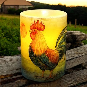 May include: A cylindrical candle with a rooster illustration. The candle has a yellow background with a colorful rooster design. The rooster is depicted with red, orange, yellow, green, and blue hues. The candle is placed on a wooden surface.
