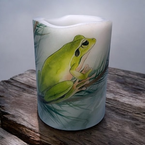 May include: A white pillar candle with a green tree frog sitting on a branch. The frog is painted in watercolor style on the candle.