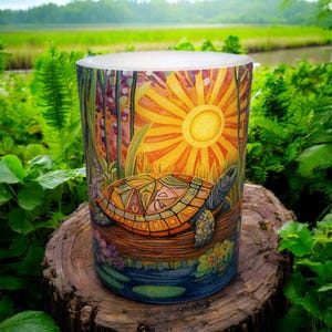 May include: A cylindrical candle holder with a colorful illustration of a turtle resting on a log in a pond, with a bright sun in the background. The candle holder is set on a tree stump, surrounded by lush green foliage.