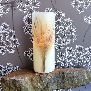 May include: A white pillar candle with a dried floral bouquet design. The bouquet features dried grasses and flowers in shades of beige, brown, and yellow.