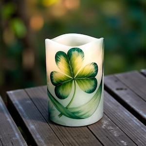 May include: A cream-colored, battery-operated candle with a green shamrock design. The shamrock has three heart-shaped leaves with yellow and green shading. The candle sits on a weathered wooden surface, with a blurred green background.