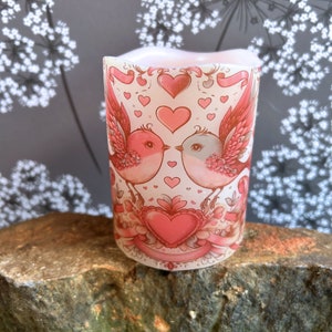 May include: A cylindrical candle with a romantic design. The candle features two birds facing each other, surrounded by hearts and ribbons in shades of pink and red. The candle is placed on a rock.